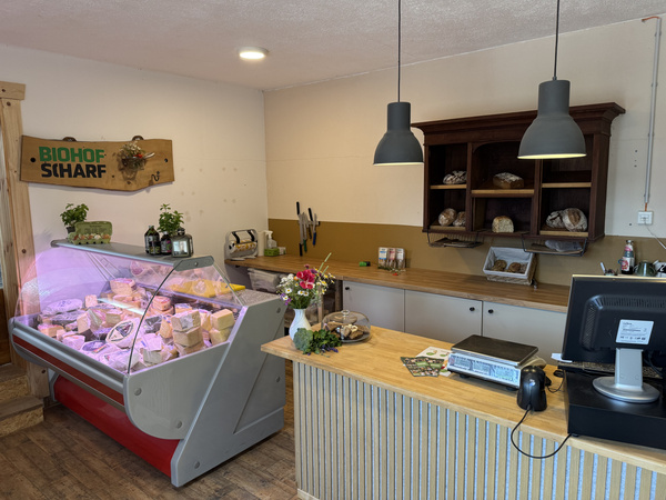 KI generiert: Einladender Hofladen mit Käsetheke, Waage, Broten und Produkten. Text: "BIOHOF SCHARF".