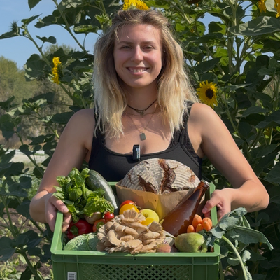 KI generiert: Eine Frau hält eine Kiste voller Obst, Gemüse und Brot vor Sonnenblumen.