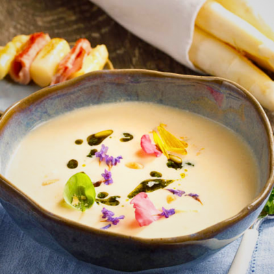 KI generiert: Eine Schüssel cremige Suppe, dekoriert mit bunten Blüten und Kräutern, neben weißem Spargel.