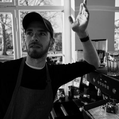 KI generiert: Das Bild zeigt eine Person in einem Café, die vor einer Espressomaschine steht. Sie trägt eine Schürze und scheint in einer entspannten Haltung zu sein.