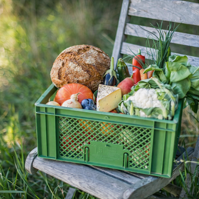 KI generiert: Grüne Kiste voller frischem Gemüse, Brot und Käse auf einem Holzstuhl im Freien.