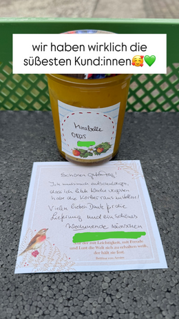 KI generiert: Ein Glas Mirabellen-Orips steht auf einer Kiste, daneben eine handgeschriebene Notiz. Text: „wir haben wirklich die süßesten Kund:innen🥰💚“