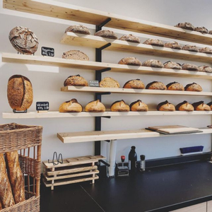 KI generiert: Das Bild zeigt eine Bäckerei mit mehreren Regalen voller verschiedener Brotsorten. Im Vordergrund sind Körbe mit Baguettes zu sehen, und darunter befinden sich Schneidebretter und Stempel.