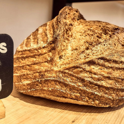 KI generiert: Das Bild zeigt ein frisch gebackenes, rustikales Brot mit einer knusprigen Kruste auf einem Holzbrett. Daneben steht ein kleines Schild, das teilweise lesbar ist.